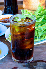 Pop water or cola in glass with ice cubes with food in the background.