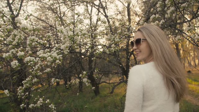 Attractive young woman in sweater, jeans is dance and spinning among blossom apple tree. Sunset, spring orchard and green field. Slow motion. Caucasian one girl, long fluttering hair. Sun glasses.