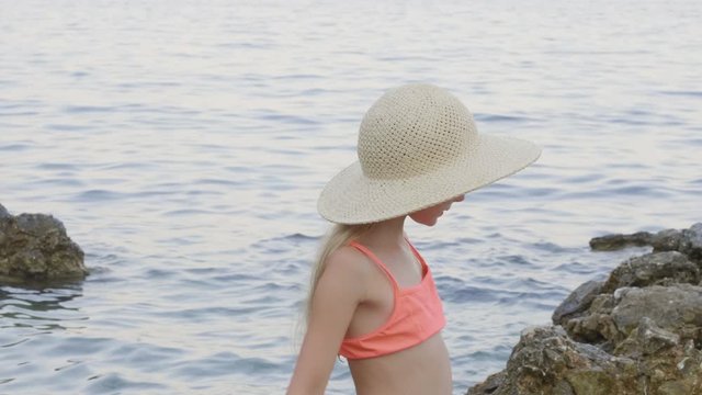 Young girl in swimsuit wearing straw hat on stony sea beach at summer vacation. Smiling teenager girl in hat on sea water on rocky beach