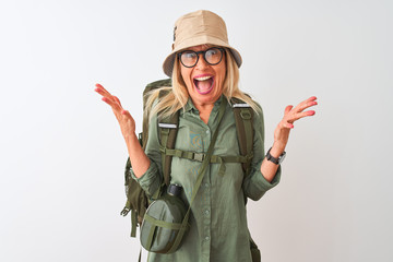 Middle age hiker woman wearing backpack hat canteen glasses over isolated white background celebrating crazy and amazed for success with arms raised and open eyes screaming excited. Winner concept