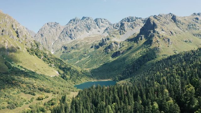 Beautiful panoramic view of Kardyvach lake surrounded high mountain ranges, deep pine fir forest on slopes, rocky cliffs and peaks; dangerous trekking route; autumn nature of Caucasus region of Russia