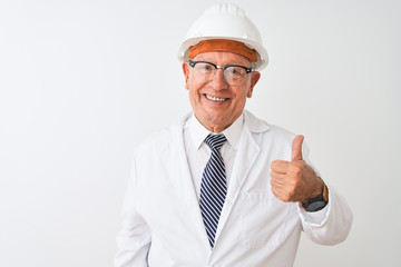 Senior grey-haired engineer man wearing coat and helmet over isolated white background doing happy thumbs up gesture with hand. Approving expression looking at the camera with showing success.
