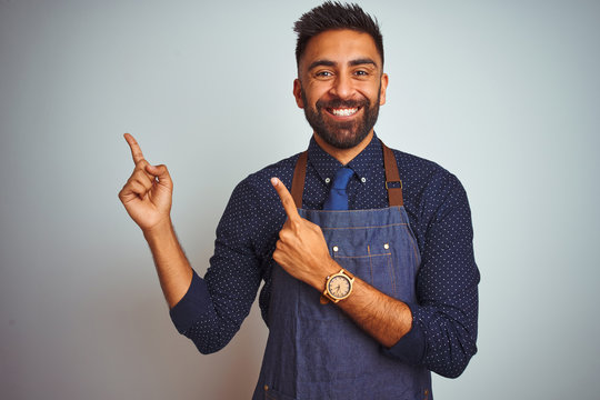 Young indian employee man wearing apron uniform standing over isolated white background smiling and looking at the camera pointing with two hands and fingers to the side.