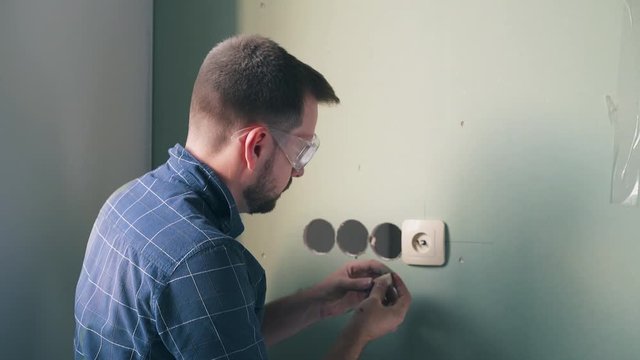 professional electrician in blue shirt installs modern lever in white plastic case on green wall and poses to camera in flat