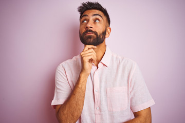 Young indian man wearing casual shirt standing over isolated pink background with hand on chin thinking about question, pensive expression. Smiling with thoughtful face. Doubt concept.