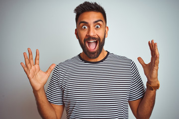 Young indian man wearing black striped t-shirt standing over isolated white background celebrating crazy and amazed for success with arms raised and open eyes screaming excited. Winner concept