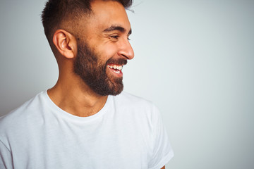 Young indian man wearing t-shirt standing over isolated white background looking away to side with smile on face, natural expression. Laughing confident.