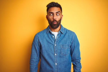 Obraz premium Young indian man wearing denim shirt standing over isolated yellow background making fish face with lips, crazy and comical gesture. Funny expression.