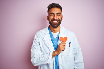 Young indian doctor man holding paper heart standing over isolated pink background with a happy...