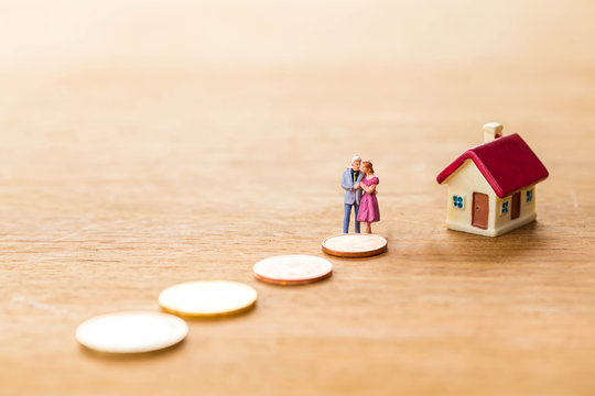 Miniature Older Couple Family Stand On Stack Coins With Tiny Home Over Wooden  Background,Image For Home And Money Management Concept.