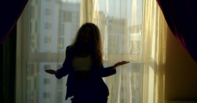 a curly-haired young beautiful girl who is standing in front of a large window, wearing a white short top, black pants and a jacket, she poses and touches her hair, makes dancing movements.