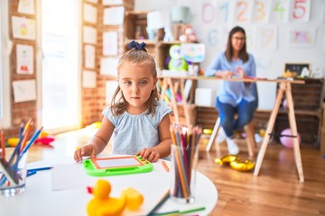 Caucasian girl kid playing and learning at playschool with female teacher. Mother and daughter at playroom around toys drawing on magnetic blackboard