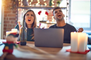 Young beautiful couple sitting using laptop around christmas decoration at home crazy and mad shouting and yelling with aggressive expression and arms raised. Frustration concept.