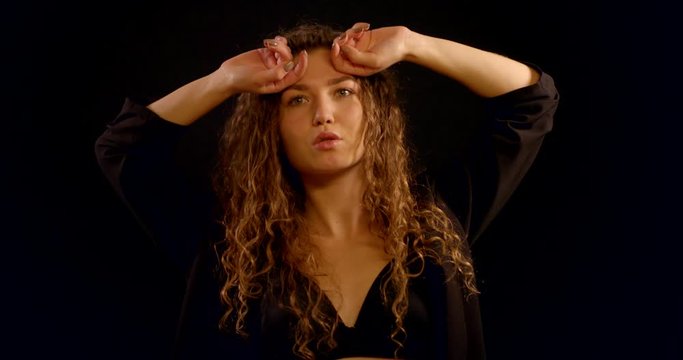 A close-up portrait of a smiling curly-haired young girl who is in the Studio on a black background, wearing a black unbuttoned jacket and a black bra, she poses and touches her hair, dances.