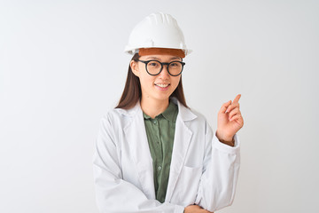 Young chinese engineer woman wearing coat helmet glasses over isolated white background with a big smile on face, pointing with hand and finger to the side looking at the camera.