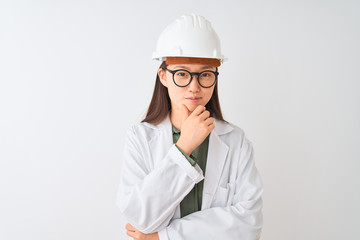 Young chinese engineer woman wearing coat helmet glasses over isolated white background looking confident at the camera with smile with crossed arms and hand raised on chin. Thinking positive.