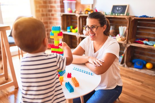 Beautiful teacher and toddler boy playing with construction blocks bulding tower at kindergarten