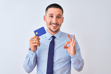 Young business man holding credit card over isolated background doing ok sign with fingers, excellent symbol