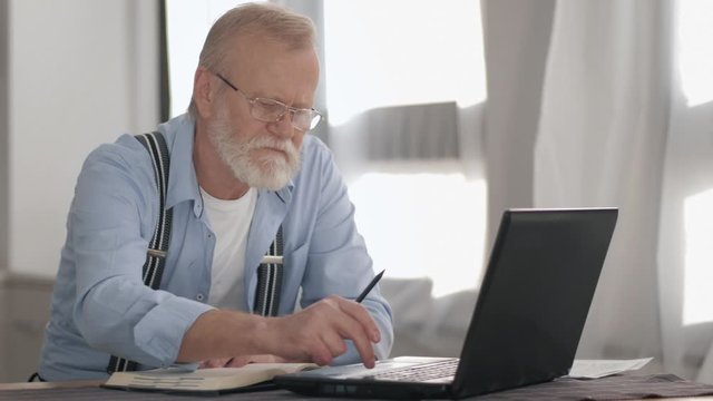 portrait of an elderly man, old male in glasses with a beard working at a computer and parses bank accounts to pay online sitting at a laptop at a table