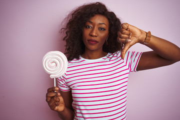 Young african american woman eating sweet candy standing over isolated pink background with angry face, negative sign showing dislike with thumbs down, rejection concept