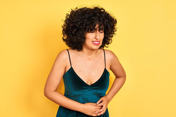 Young arab woman with curly hair wearing elegant dress over isolated yellow background with hand on stomach because indigestion, painful illness feeling unwell. Ache concept.