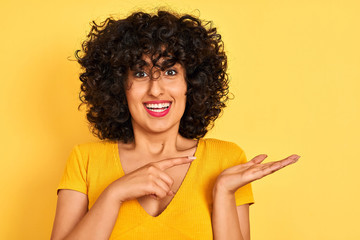 Young arab woman with curly hair wearing t-shirt standing over isolated yellow background very happy pointing with hand and finger