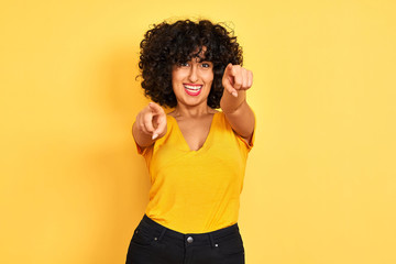 Young arab woman with curly hair wearing t-shirt standing over isolated yellow background pointing to you and the camera with fingers, smiling positive and cheerful