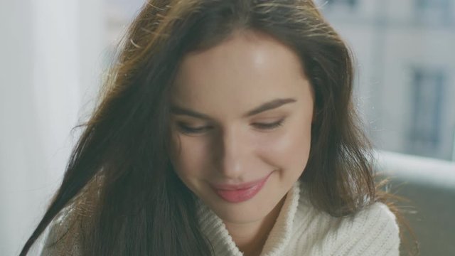 Portrait of Young Woman Smiling Charmingly while Sitting on a Cauch, Brushes Her Lush Hair Away with Flirtatious Gesture. Girl Wearing White Knitted Sweater Relaxes on a Couch in Her Cozy Apartment