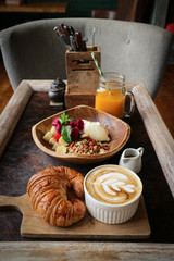 A cup of coffee with latte art on top, butter  croissant and granola with fresh tropical fruits and yogurt on table in caffee. Concept of healthy breakfast