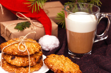 Closeup of Christmas crispy oatmeal cookies, transparent mug of coffee with milk or cappuccino, gift boxes wrapped in kraft paper, hat forgotten by Santa Claus on a brown background. Holidays concept