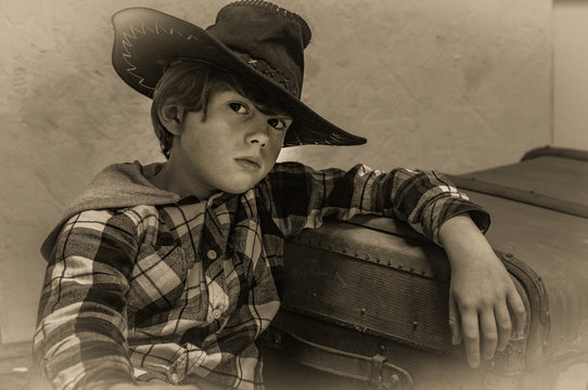 Boy model in a cowboy hat and a plaid shirt of blond with long hair on a background of a suitcase in retro style. Vintage style, sepia. Art noise