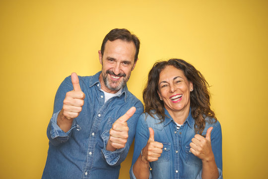 Beautiful middle age couple together standing over isolated yellow background success sign doing positive gesture with hand, thumbs up smiling and happy. Cheerful expression and winner gesture.