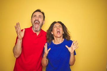 Fototapeta premium Beautiful middle age couple together standing over isolated yellow background crazy and mad shouting and yelling with aggressive expression and arms raised. Frustration concept.