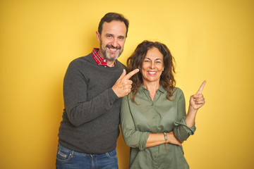 Beautiful middle age couple over isolated yellow background with a big smile on face, pointing with hand and finger to the side looking at the camera.
