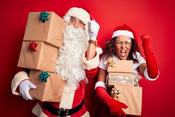 Middle age couple wearing Santa costume holding tower of gifts over isolated red background angry and mad raising fist frustrated and furious while shouting with anger. Rage and aggressive concept.