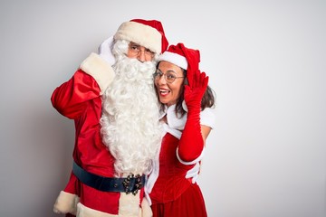 Middle age couple wearing Santa costume hugging over isolated white background smiling with hand over ear listening an hearing to rumor or gossip. Deafness concept.