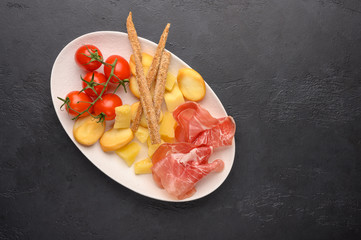 Traditional Italian food is grissini bread with prosciutto, cheese and tomatoes with herbs on a plate on a dark background. Healthy style. Copy space
