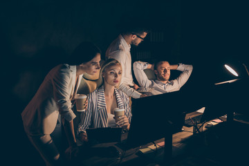 Photo of four business people partners working late night drink coffee watch graphics stats seriously reading startup project report workaholics formalwear dark office indoors