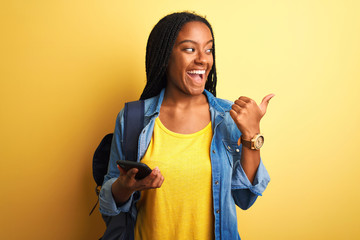 African american student woman using smartphone standing over isolated yellow background pointing and showing with thumb up to the side with happy face smiling
