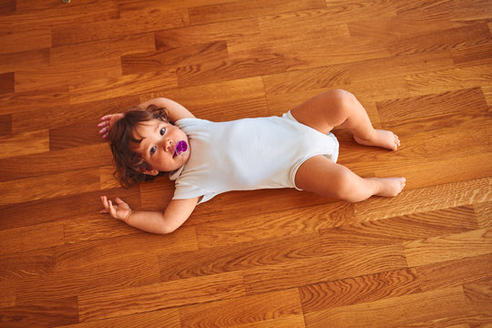 Beautiful toddler child girl wearing white bodysuit lying down on the floor using pacifier