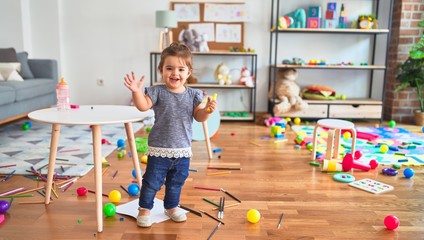 Beautiful toddler holding yellow pen standing around lots of toys smiling at kindergarten