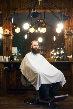 Full length portrait of bearded man overgrown sitting in retro chair before haircutting in modern barber shop. Big mirror at professional equipped workplace illuminated by many lamps