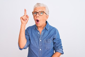 Senior grey-haired woman wearing denim shirt and glasses over isolated white background pointing finger up with successful idea. Exited and happy. Number one.