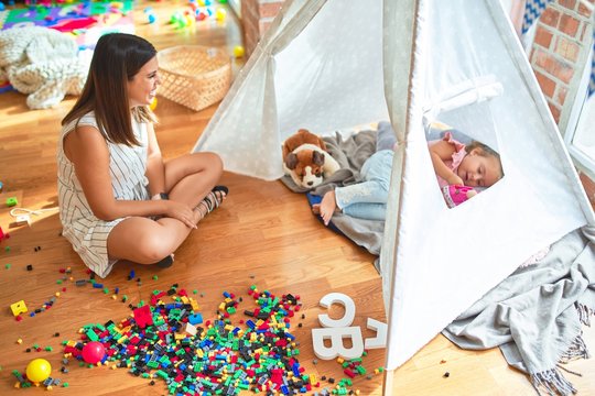 Beautiful teacher and blond toddler girl playing with dolls inside tipi at kindergarten