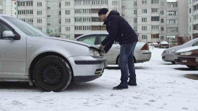 Man trying and can not open hood of his car in winter in parking lot of yard. Bonnet froze due to cold weather. Malfunction or problem with auto. Breakdown. One wants to charge the discharged battery