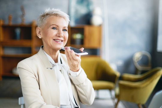 People, communication, networking and modern electronic devices concept. Indoor shot of short haired European female in stylish suit recording voice message holding smart phone at her mouth, speaking