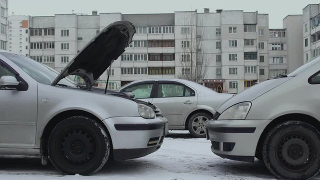 Car pulls up to broken auto to help and to charge discharged automotive battery. Hood raised up in winter daytime in parking lot of yard. One wants to charge discharged battery using cables. Part 1