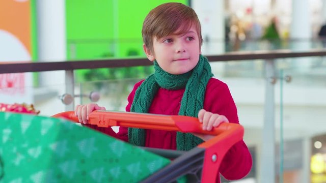Funny boy walk in the shopping mall with Christmas present boxes looks to side in cart child family attractive teenager beautiful children cheerful happiness holiday kid snile childhood slow motion