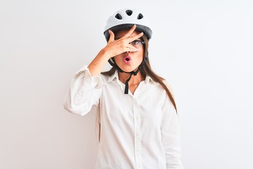 Beautiful businesswoman wearing glasses and bike helmet over isolated white background peeking in shock covering face and eyes with hand, looking through fingers with embarrassed expression.