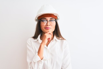 Young beautiful architect woman wearing helmet and glasses over isolated white background looking confident at the camera with smile with crossed arms and hand raised on chin. Thinking positive.